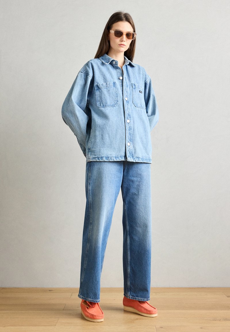 Camisa de mezclilla oversize con dos bolsillos en el pecho, combinada con pantalones vaqueros anchos de color azul claro y zapatillas naranjas brillantes. Gafas de sol marrones.