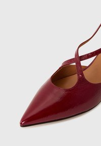 Chaussure à bout pointu en cuir verni bordeaux brillant, présentant un design découpé et deux brides croisées. Texture lisse avec une semelle plate.