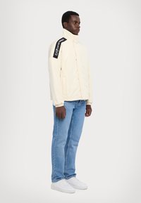 Cremefarvet jakke med høj krave, prydet med sort logo-detalje på skulderen, parret med blå jeans og hvide sneakers. Glat tekstur.