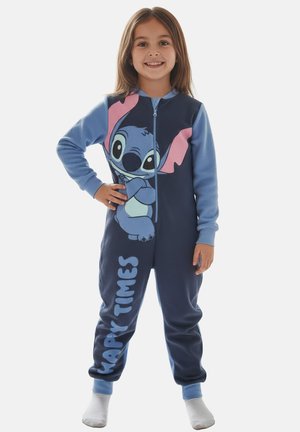 Smiling girl standing with hand on hip, wearing blue onesie featuring cartoon character Stitch and "Happy Times" text, white socks.
