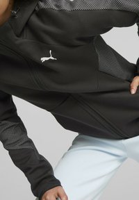 Czarna bluza z zamkiem błyskawicznym, z teksturowanymi szarymi panelami i białym logo na klatce piersiowej. Wykonana z gładkiego, elastycznego materiału; posiada kaptur i dopasowany krój.