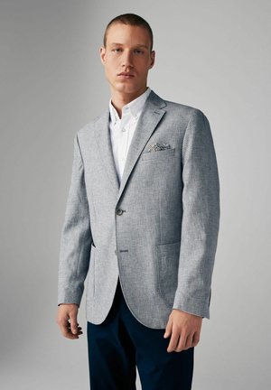 Blazer gris claro con dos botones, solapas de muesca y tela texturizada. Presenta un pañuelo de bolsillo con un patrón floral. Combinado con una camisa blanca.