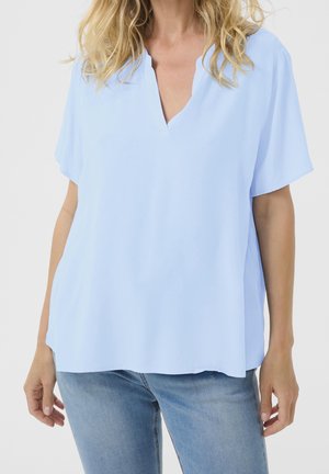 Mujer con blusa azul claro de manga corta con escote en V y vaqueros azules, con cabello rubio suelto y pose relajada sobre fondo liso.