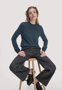 Donkerblauwe trui met een ronde hals, gecombineerd met grijze broekspijpen met streepjes en een wijde pasvorm. Model zit op een houten kruk en draagt zwarte franjeschoenen.