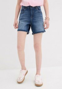 Shorts di jeans a vita alta di blu medio con risvolti, dotati di tasche e una texture liscia. Abbinati a scarpe sportive bianche e rosa.