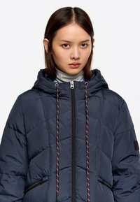 Młoda kobieta ubrana w granatową pikowaną kurtkę z kapturem, z bordowymi troczkami, oraz jasnoszary golf pod spodem.