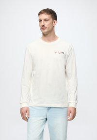 Camisa crema de manga larga con un escote redondo y puños. Presenta un pequeño diseño bordado en el pecho. Se lleva con jeans azul claro.