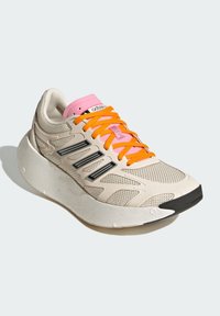 Beiger Sportschuh mit einem strukturierten Mesh-Obermaterial, rosa Zunge, orangen Schnürsenkeln und einer klobigen weißen Sohle mit schwarzem Akzent vorne.