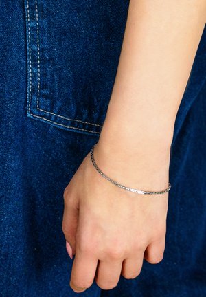 Poignet avec bracelet à chaîne argentée fine reposant contre un tissu en denim bleu foncé avec poche visible et détails de couture.