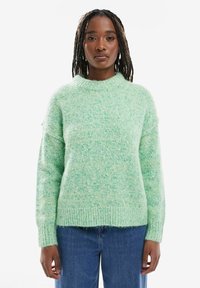 Femme aux cheveux tressés portant un pull en tricot vert clair et un jean bleu, debout devant un fond blanc.