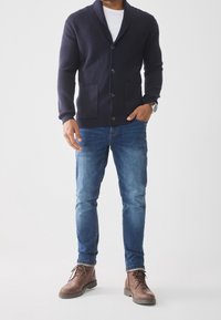 Granatowy prążkowany kardigan z kołnierzem typu szal, pięcioma guzikami i dwiema przednimi kieszeniami. Noszony na białej koszulce i w zestawieniu z niebieskimi dżinsami oraz brązowymi butami.