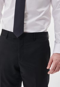 Black formal trousers with a smooth texture, flat front, and straight-cut design. Paired with a white dress shirt and dark tie.