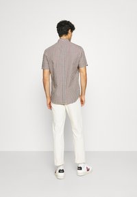 Camicia a maniche corte a quadri in rosso, blu e bianco; vista posteriore che mostra un design aderente con colletto, abbinata a pantaloni chiari e sneakers.