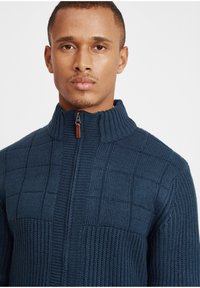Sötétkék kötött pulóver magas gallérral és cipzáras záródással, amely textúrázott négyzetrács mintával és bordás díszítésekkel rendelkezik az oldalakon.
