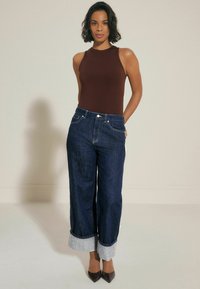 Bruine mouwloze top gecombineerd met high-waisted donkerblauwe spijkerbroek met opvouwbare cuffs, aangevuld met donkere, puntige hoge hakken.
