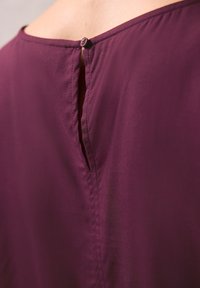 Bordeaux bluse i stof med en glat tekstur, prydet med en diskret knaplukning bagpå og en slids for at tilføje detalje nær halsudskæringen.