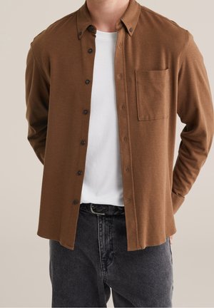 Mann trägt ein offenes braunes Hemd über einem weißen T-Shirt mit schwarzem Gürtel und dunklen Jeans, Hände hinter dem Rücken, neutraler Hintergrund.