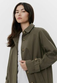 Trench coat verde oliva con colletto, grandi bottoni e vestibilità rilassata, indossato sopra una maglia bianca. Tessuto morbido con una sottile trama.
