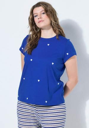 Frau mit welligem Haar, die ein blaues T-Shirt mit kleinen weißen Herzen und gestreifte Hose trägt, steht mit den Händen hinter dem Rücken vor einer weißen Wand.