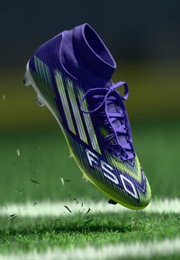 Crampon de fotbal violet și verde lămâie cu guler înalt, având dungi texturate, accente albe și șireturi vizibile.