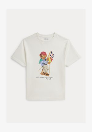 Biały t-shirt z krótkim rękawem, na którym znajduje się niedźwiedź w czerwonej czapce, w paskowanej koszuli i khakich spodniach, trzymający na huśtawce flagi żeglarskie.