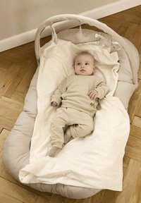 Wieg met een zachte, gestoffeerde grijze omranding, voorzien van een witte deken en een gestructureerde basis. Een baby in tan kleding ligt binnenin.