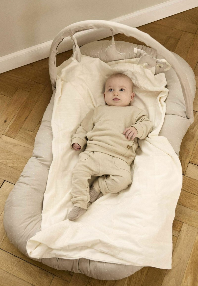Wieg met een zachte, gestoffeerde grijze omranding, voorzien van een witte deken en een gestructureerde basis. Een baby in tan kleding ligt binnenin.