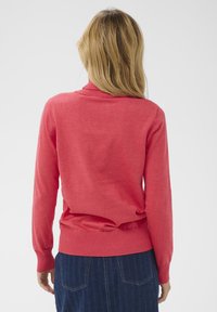 Pull-over corail avec col et ourlet en côte, manches longues et texture douce. Porté avec une jupe en denim foncé.