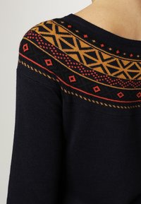 Marineblauer Pullover mit geometrischem Muster in Goldgelb und Rot über der Schulter, mit einer weichen Textur und mittelschwerer Strickart.