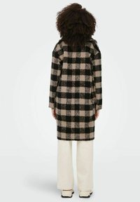 Long manteau à carreaux avec un motif noir et beige, confectionné en tissu texturé. Il possède une coupe décontractée et est porté avec un pantalon de couleur claire.