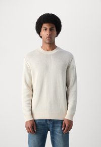Suéter de punto color beige claro con cuello redondo y puños acanalados, combinado con jeans de mezclilla azul. Textura suave, corte clásico.