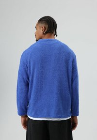 Blauwe, getextureerde fleece trui met lange mouwen en een ronde hals. Gedragen over een wit overhemd, weergegeven van achteren met een losse pasvorm.