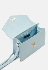 Ljusblå rektangulär axelväska med guldspärr och hörnaccenter, öppen lock som avslöjar ett dragkedjeförsett innerfack och tygfoder.