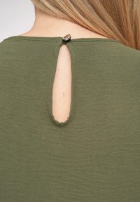 Olivegrünes Top mit einem Schlüsselaugen-Ausschnitt hinten und einer Metallknopfverschluss, aus weichem, strukturiertem Stoff mit glatter Oberfläche gefertigt.