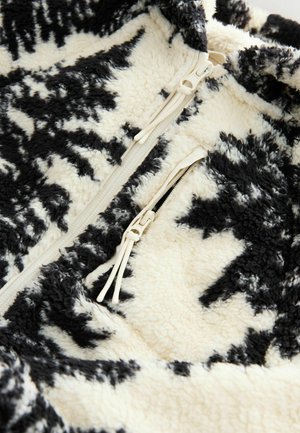 Fleece jacket in cream and black, featuring a soft, textured surface with a geometric pattern, front zipper, and side pocket with zipper.