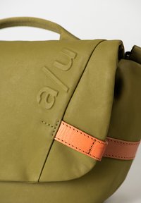 Borsa verde oliva con una finitura testurizzata, dotata di una tracolla in pelle arancione e un marchio impresso. Chiusura con zip e forma morbida e arrotondata.