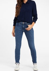 Ciemnoniebieska, prążkowana koszula z guzikami, z kieszonką na piersi, zestawiona z jasnoniebieskimi, dopasowanymi dżinsami z podwiniętymi mankietami. Całość uzupełniają białe sneakersy.