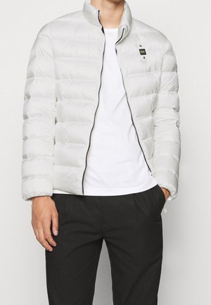 Mężczyzna ubraný w jasnoszarą pikowaną kurtkę puchową na białej koszulce i czarnych spodniach, stojący z jedną dłonią w kieszeni.