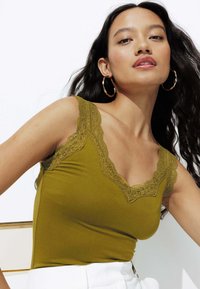 Débardeur vert olive avec un col en V bordé de dentelle et des bretelles réglables. Tissu lisse et extensible. Le mannequin porte de grandes boucles d'oreilles.