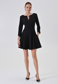 Schwarzes A-Linie-Kleid mit Dreiviertelärmeln, Schlüsselloch-Ausschnitt und einem dekorativen Broschendetail. Glatter Stoff mit einem ausgestellten Rock.