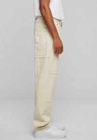 Beige cargo bukser laget av bomull med lapper, sidelommer og en hvit logo; sammen med hvite joggesko.