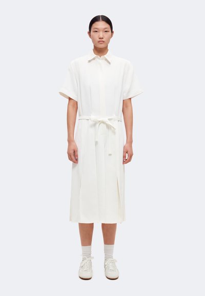 Femme debout face à nous, portant une robe chemise blanche mi-longue à manches courtes, taille nouée, chaussettes blanches et baskets blanches.