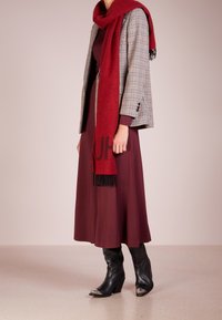 Červená šála s třásněmi, blazer v houndstooth vzoru, vínové šaty a černé boty se stříbrnými akcenty. Vrstvené textury a kontrastní barvy.
