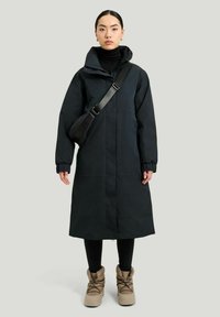 Svart huva lång kappa med dragkedja fram, elastiska ärmar, bärs över en svart polotröja och kombineras med beige stövlar. Slät tygtextur.