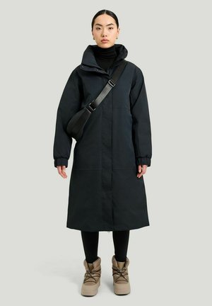 Sort hættefrakke med lang længde og frontlynlås, elastiske manchetter, båret over en sort turtleneck og parret med brune støvler. Glat stoftekstur.