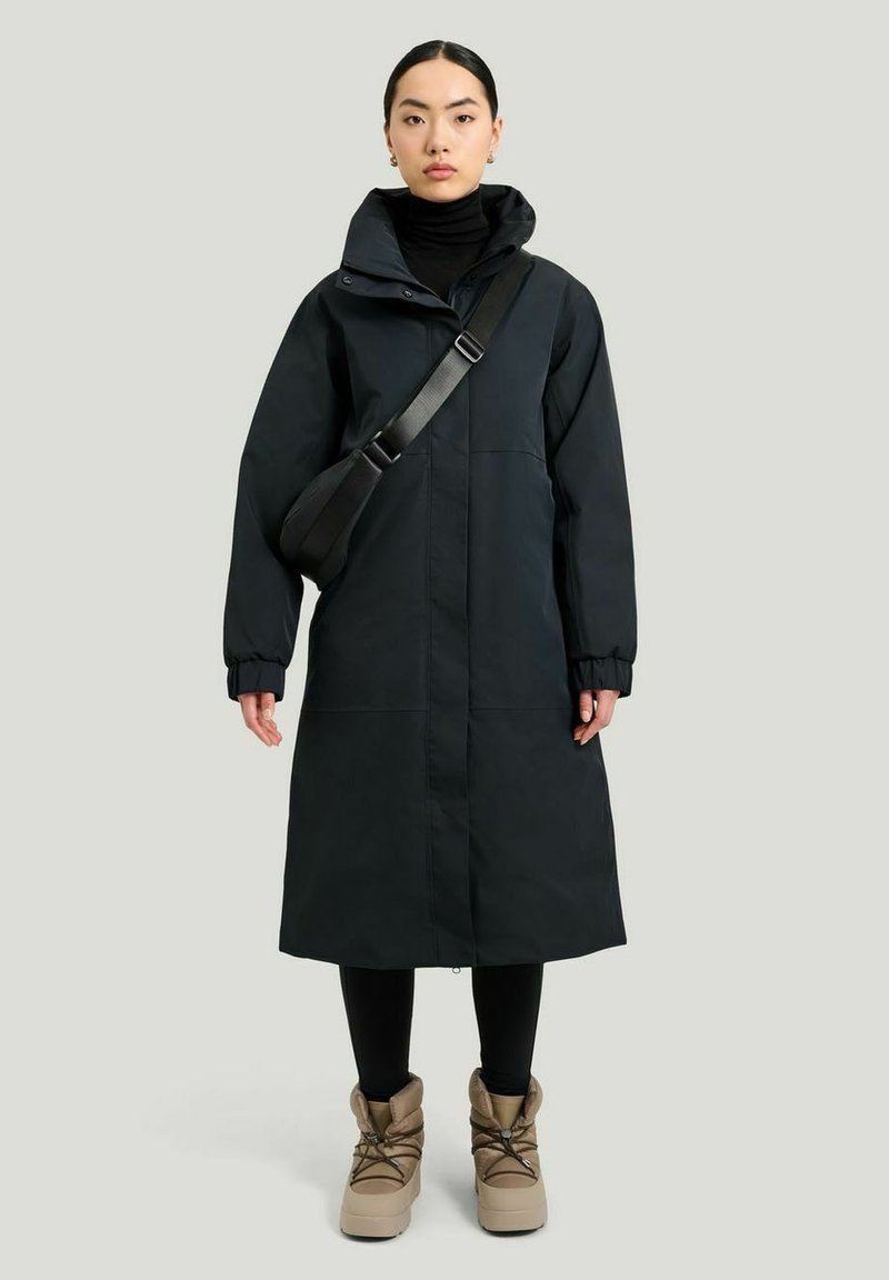 Svart huva lång kappa med dragkedja fram, elastiska ärmar, bärs över en svart polotröja och kombineras med beige stövlar. Slät tygtextur.