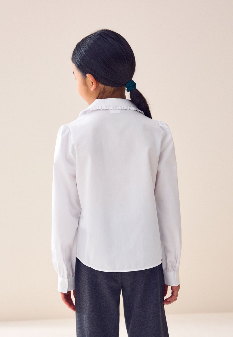 Enfant aux cheveux foncés attachés en queue de cheval, portant une chemise blanche à manches longues et un pantalon foncé, debout de dos sur un fond uni.