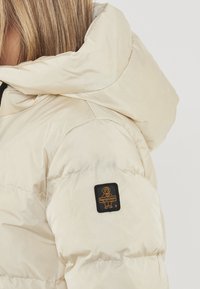 Giacca puffer beige con colletto alto e un marchio in tessuto sulla manica. Texture liscia con design trapuntato e finitura lucida.