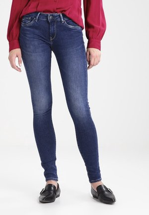 Jeans skinny azul oscuro de denim, con un diseño clásico de cinco bolsillos y un ligero desgarro en las rodillas. Combinados con una blusa roja.