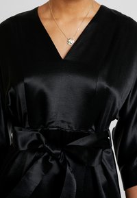 Robe en satin noir avec un décolleté en V et des manches courtes. Elle présente une taille cintrée avec un grand nœud dans le dos, offrant une texture lisse.